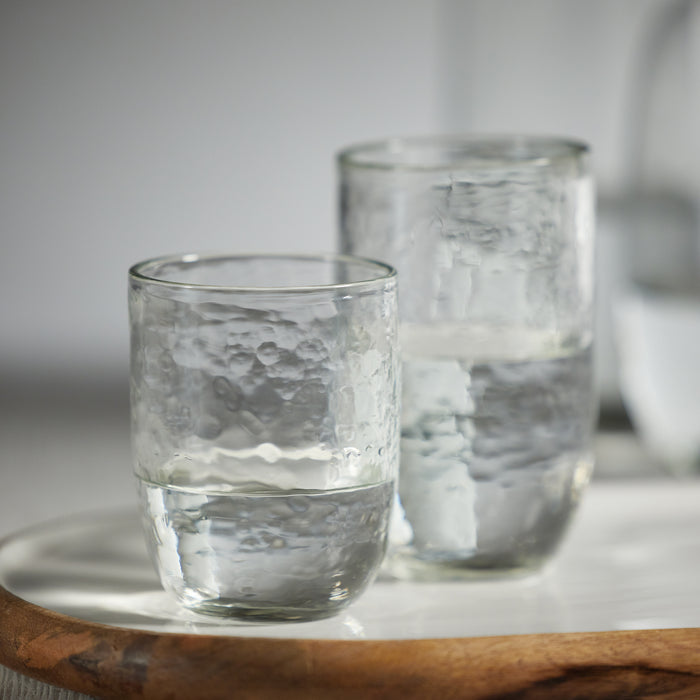 Set of four clear hammered highball glasses, textured glass with two-tone shading, on a white surface with a wooden tray