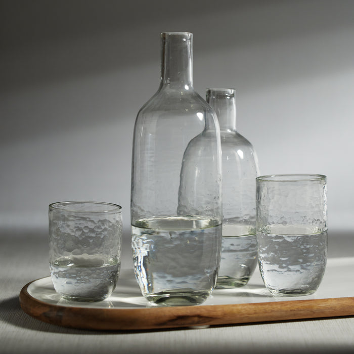 Clear glass hammered decanter with matching bottle and two small hammered glasses, all on a wooden tray against a neutral