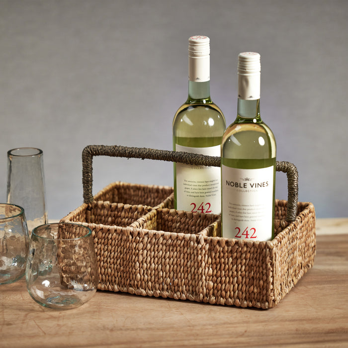 A seagrass drinkware caddy containing two bottles of wine, with divided compartments for organizing cutlery or napkins. The caddy is placed on a wooden surface with glasses and other dishes in the background.