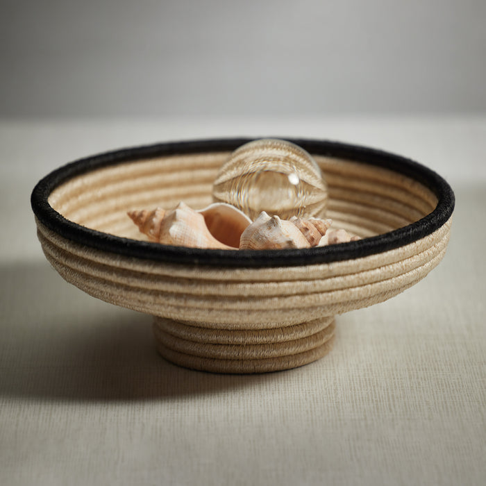 Coiled natural abaca footed bowl with black rim, filled with seashells and a glass sphere, neutral backdrop.