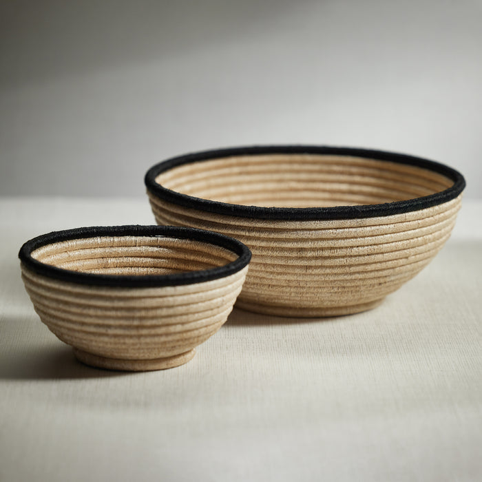 Two handwoven abaca fiber bowls on a neutral background, one large with a beige hue and a black colored rim, and one small with a beige hue and a black colored rim.