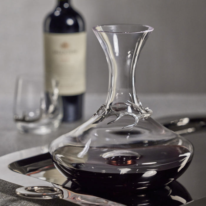 Clear glass wine decanter with a wide base and slender neck featuring pewter antler accent; dark wine inside; wine glass