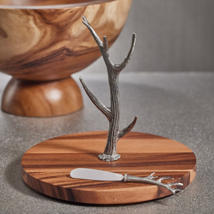 Wood round board with a silver antler-shaped cheese knife stand; metal antler stem and small blade on board.