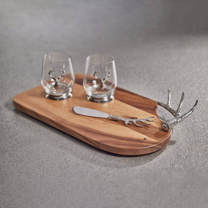 Wood serving board with pewter antler-handle cheese knife and two small glass cups featuring antler designs, neutral gray