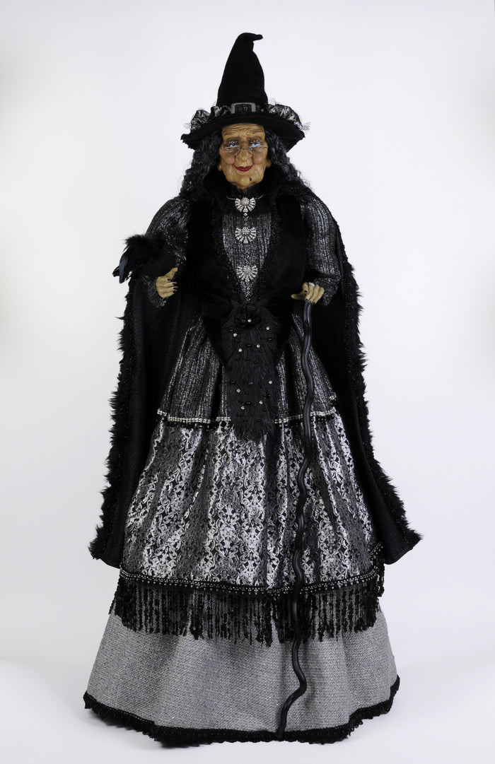 Costume figure of a witch with a black hat, cloak, and silver dress on a white background