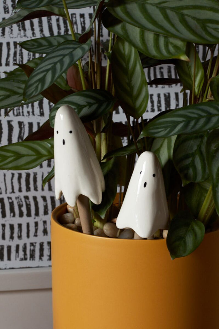 Two white ghost figurines in a potted plant with a patterned wall in the background.