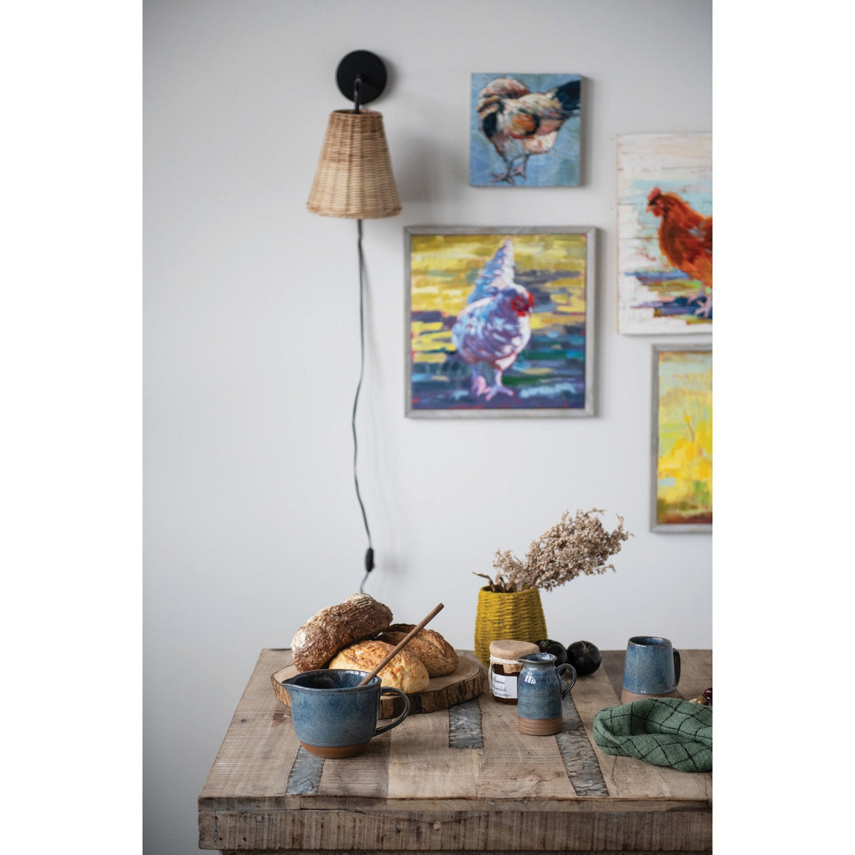 Table with bread and mugs, wall sconce, and framed rooster artwork on a light gray wall.