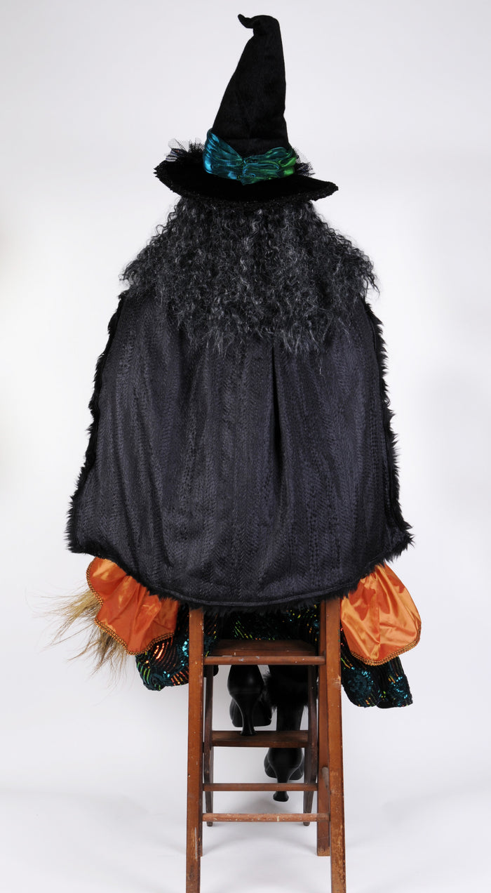 Halloween witch costume with black cape, hat, and orange accents on a wooden stool against a white background.
