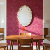 Decorative Venetian glass gold mirror inside of a dining room scene with a mid-century modern dinner wood table, a clear glass vase with one orange flower inside of it, and a magenta paisley wallpaper.
