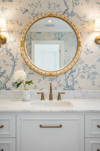gold decorative rim mirror inside of a bathroom with a marble white vanity with matte gold fixtures, a white marble cup with one white flower inside of it, and a blue printed chinoiserie wallpaper.