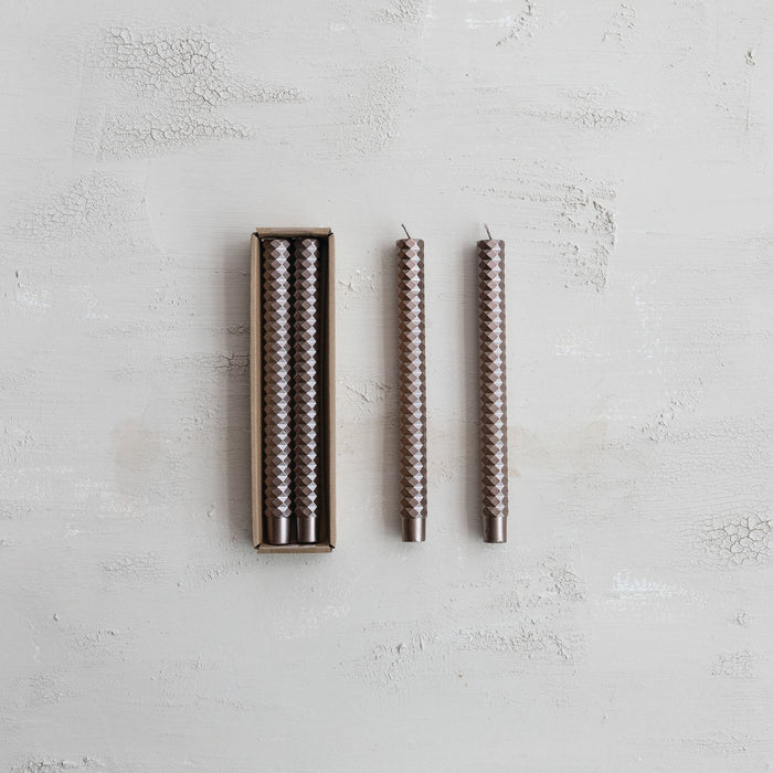 Two brown faceted taper candles with a hexagonal pattern; one in a box and three upright on a light gray textured surface.