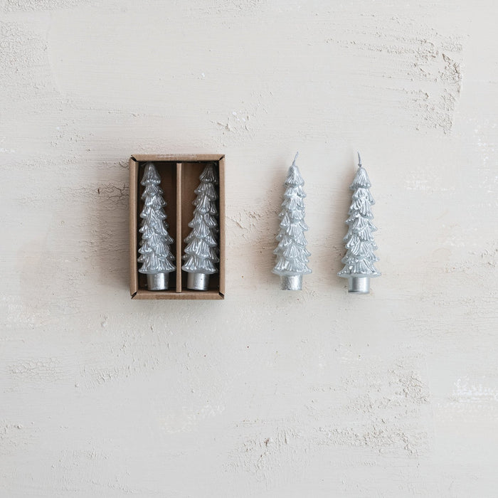 Two silver-toned tapered Christmas tree candles with textured branches, one in a brown box and two outside on a light