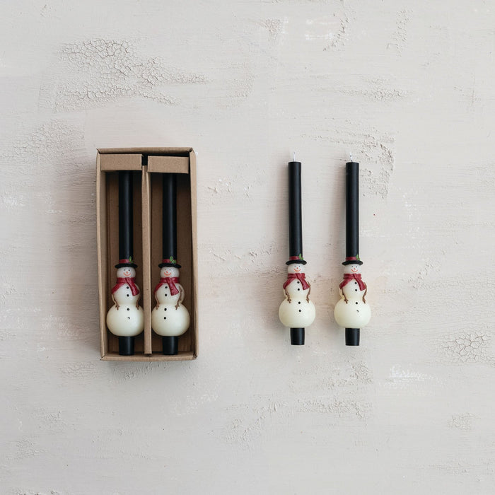Two black taper candles with snowman topper, packaged in a brown box; two more standalone candles beside, white snowmen with